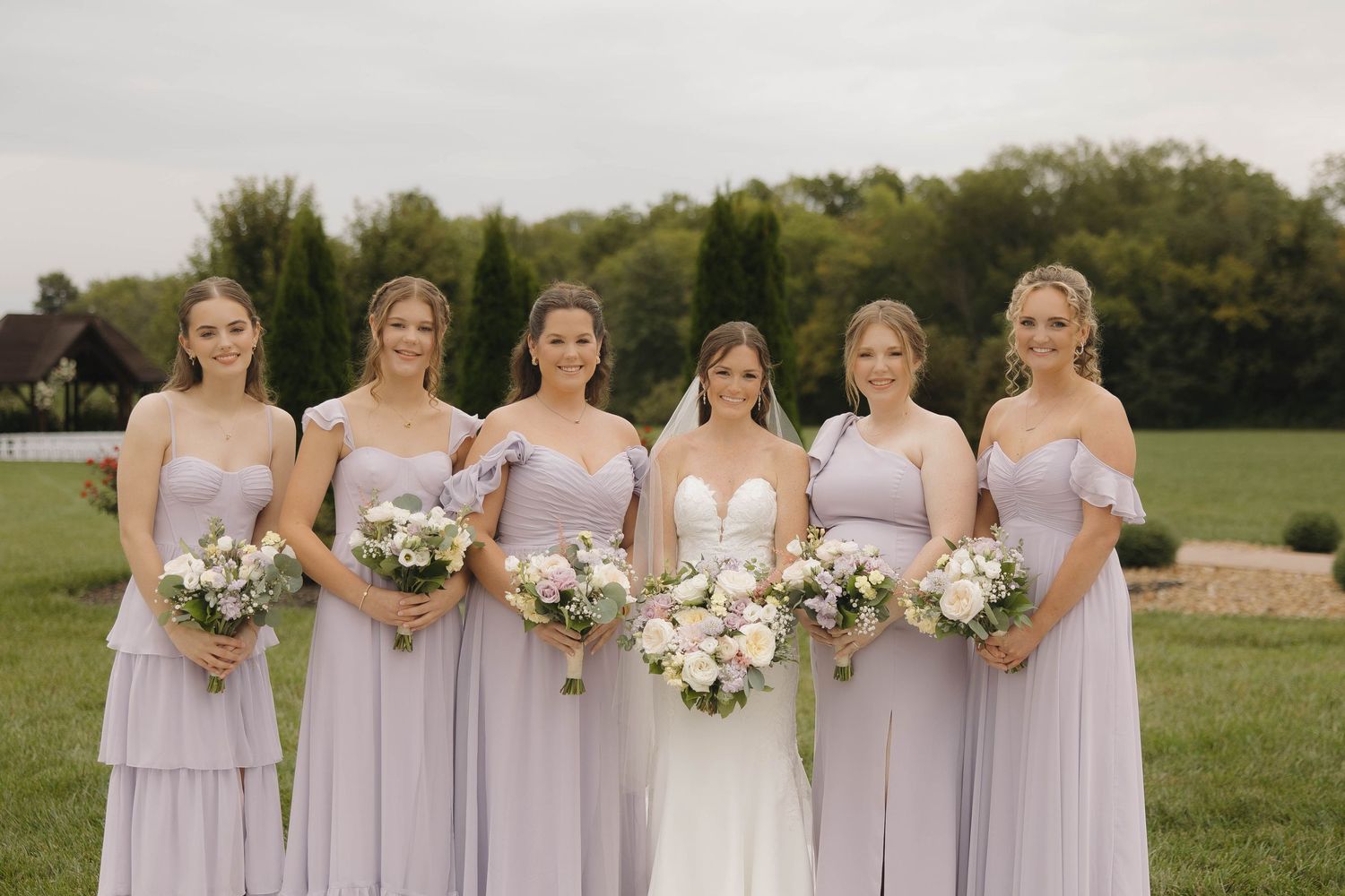 Bridesmaids in lavender dresses hold white bouquets while standing in a row outdoors.