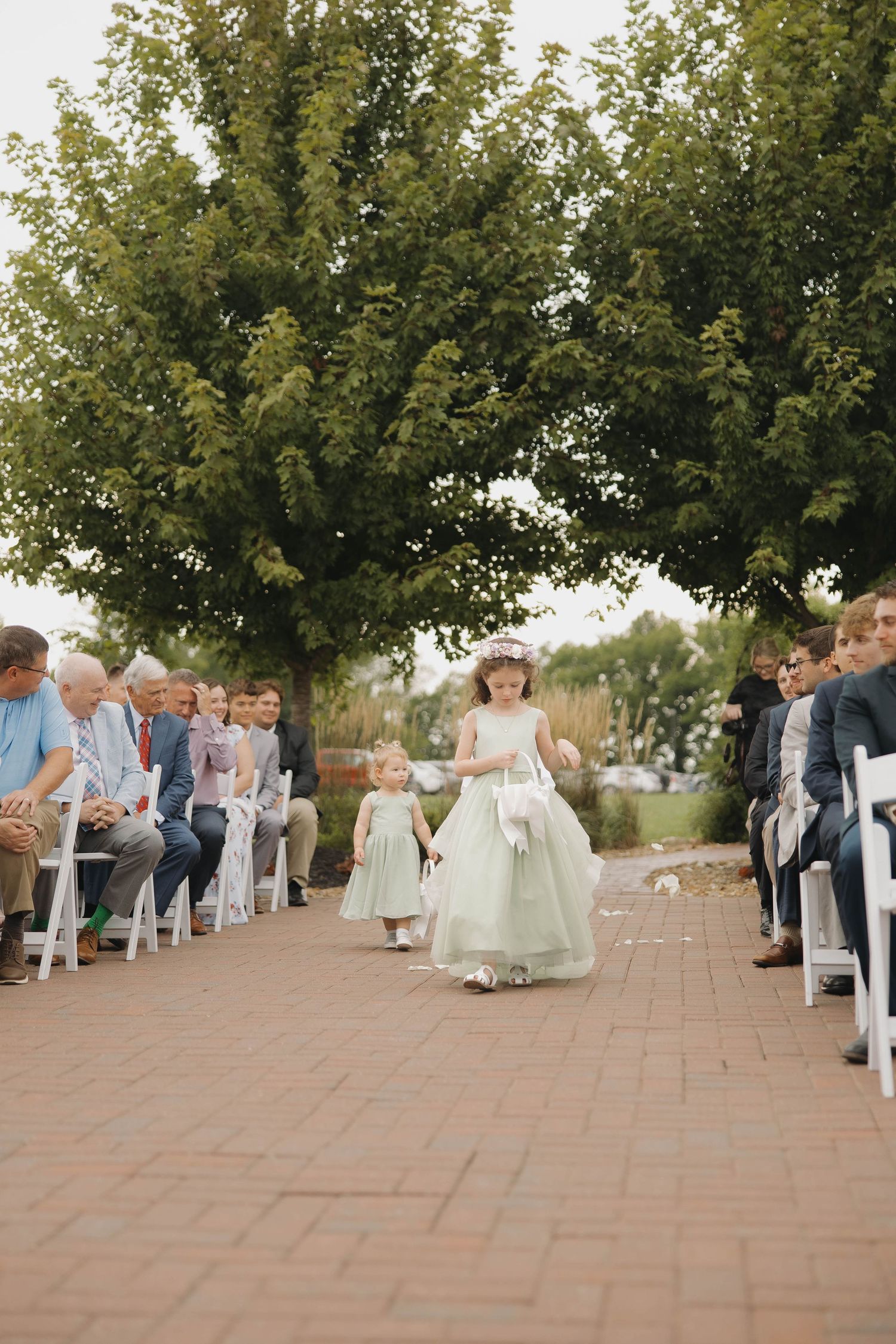 Guests lined up outdoors for a wedding ceremony procession with trees and brick pavers.