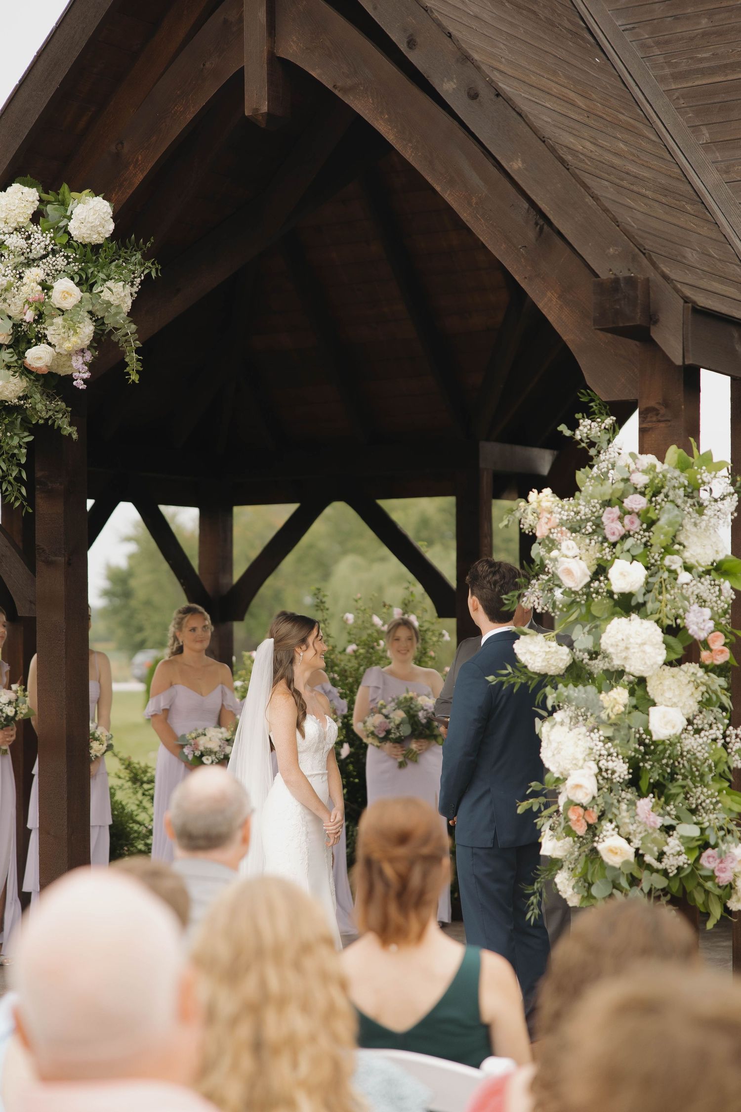 Rustic wooden pavilion decorated with white floral arrangements hosts an outdoor wedding ceremony.