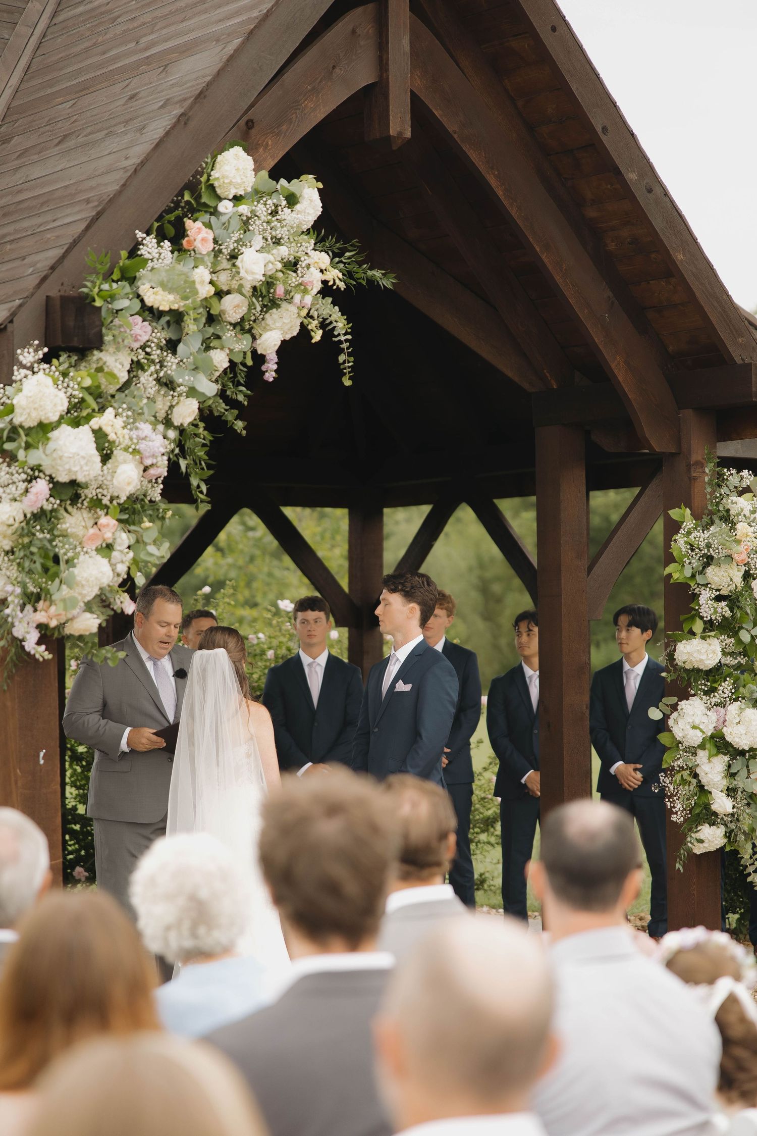 Wedding ceremony view from the back shows guests watching proceedings under a decorated wooden pavilion.