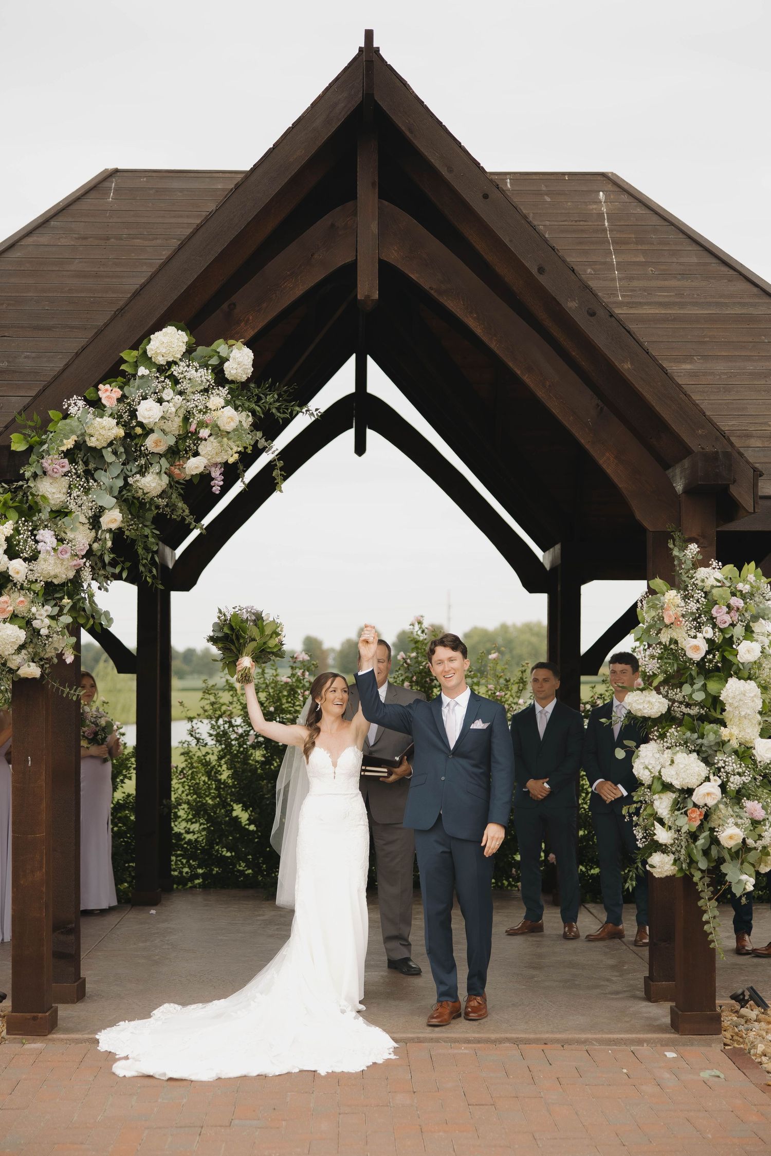 Wedding party stands under rustic wooden pergola adorned with elegant white floral arrangements and greenery.