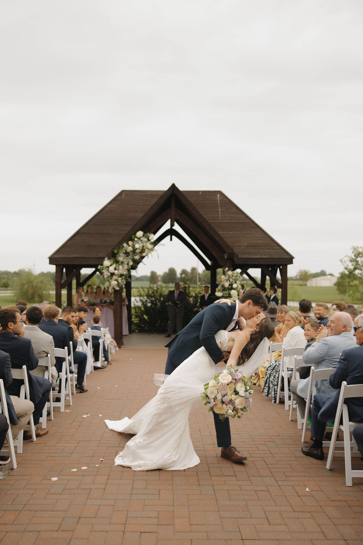 Wedding ceremony setup under a wooden pavilion with white chairs and floral decorations.