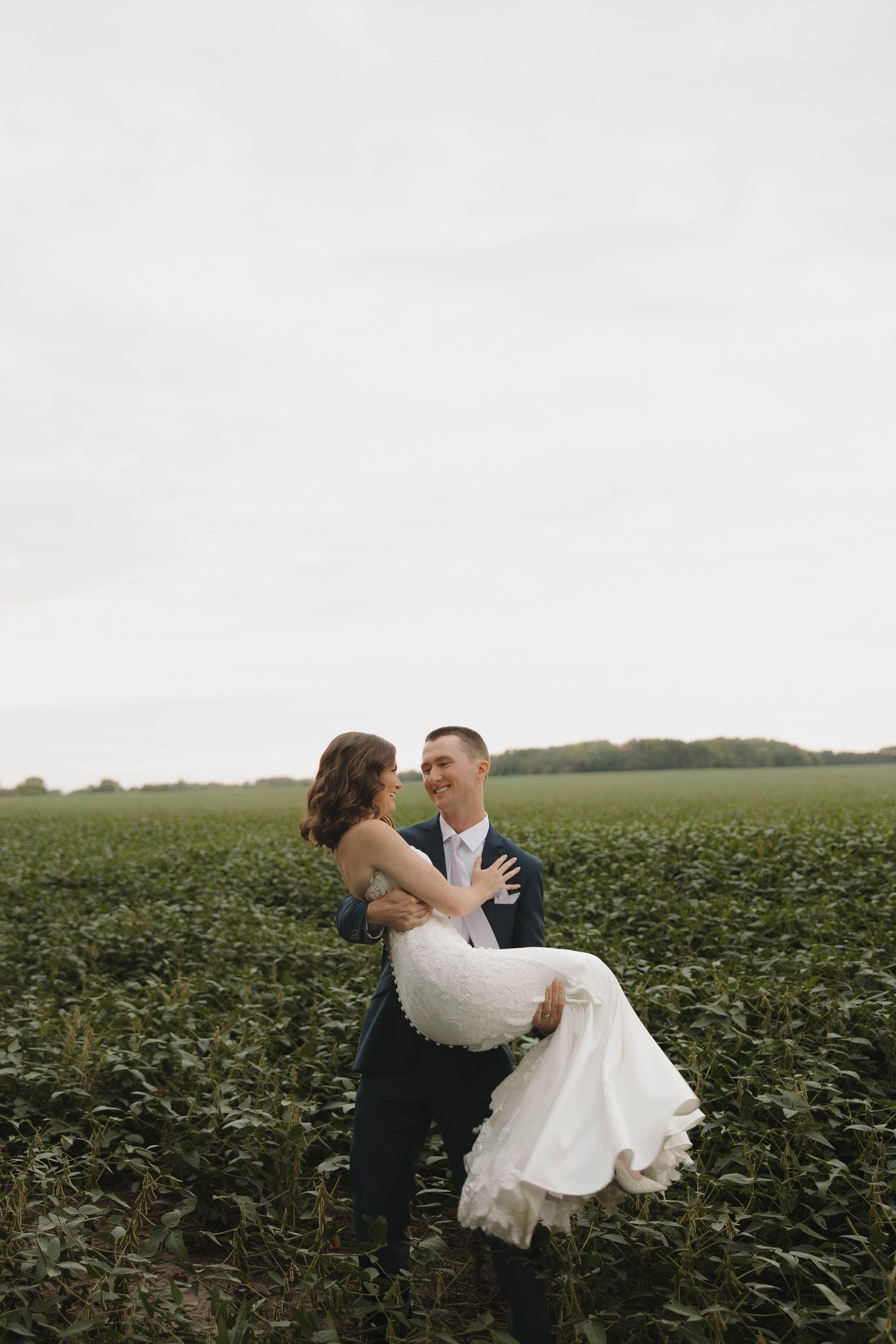 Romantic embrace in a lush green soybean field as wind blows through a white wedding dress on an overcast day.