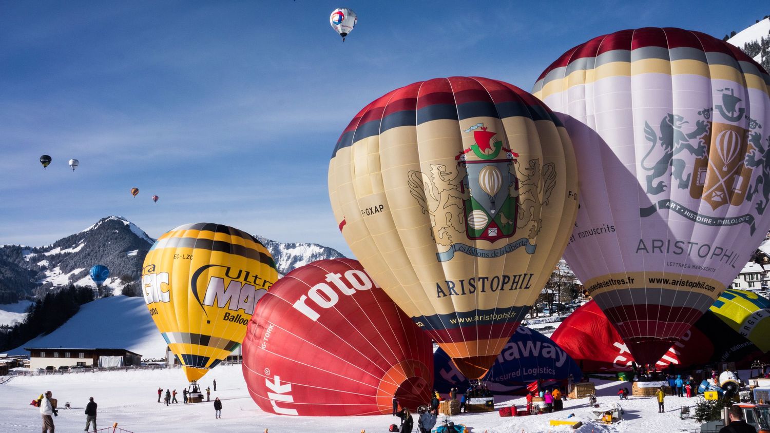 Colorful hot air balloons float against blue sky during winter festival with snow-covered mountains in background.