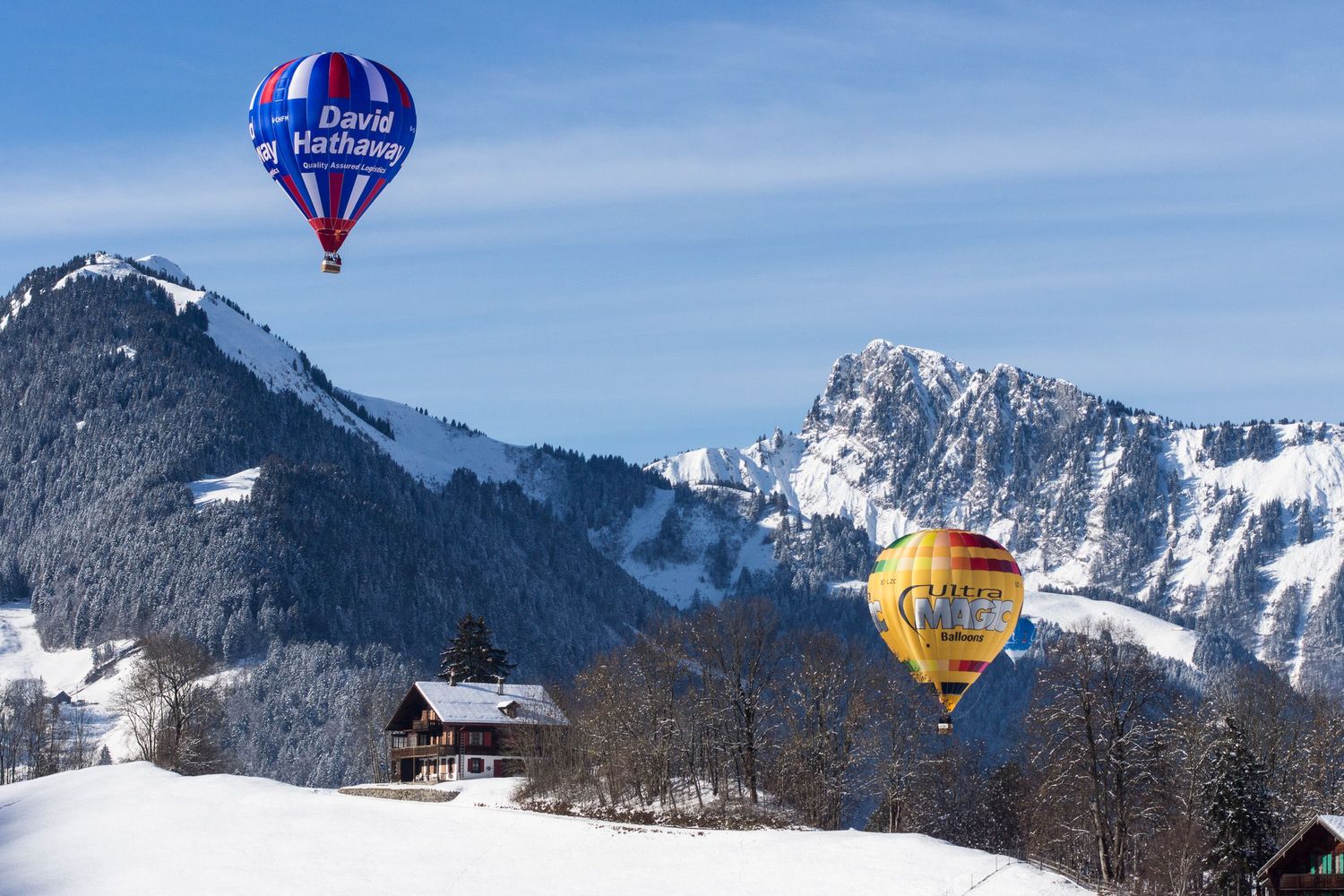Hot air balloons float above snow-covered mountains against a bright blue sky during a winter festival.