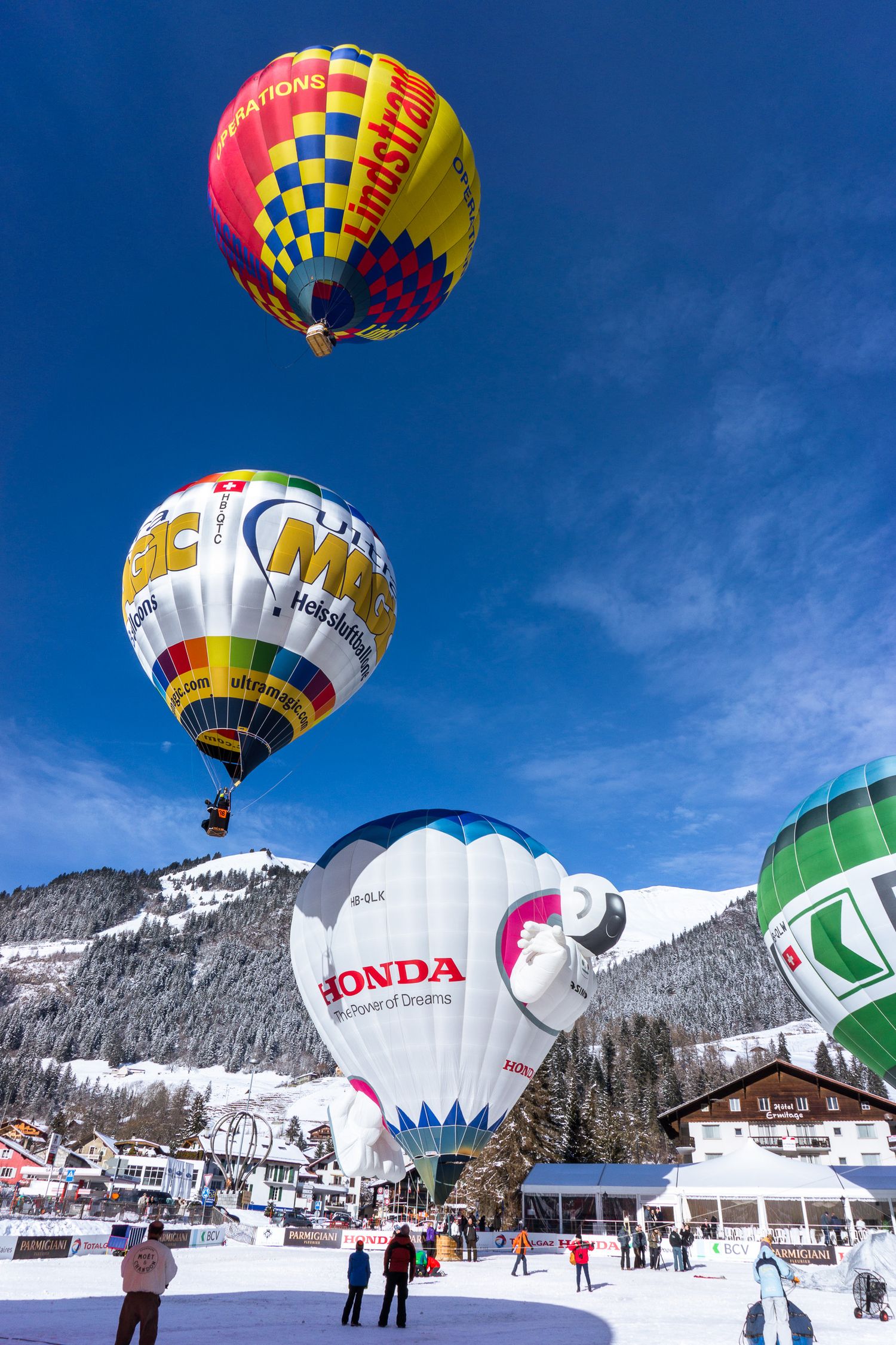 Colorful hot air balloons float against blue sky over snowy mountain landscape during winter festival in Alps.