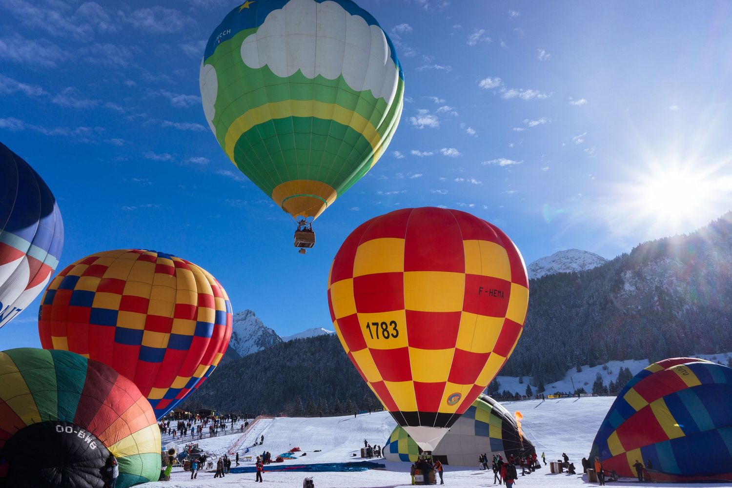 Colorful hot air balloons float above snowy mountains against a bright blue sky during a winter festival.
