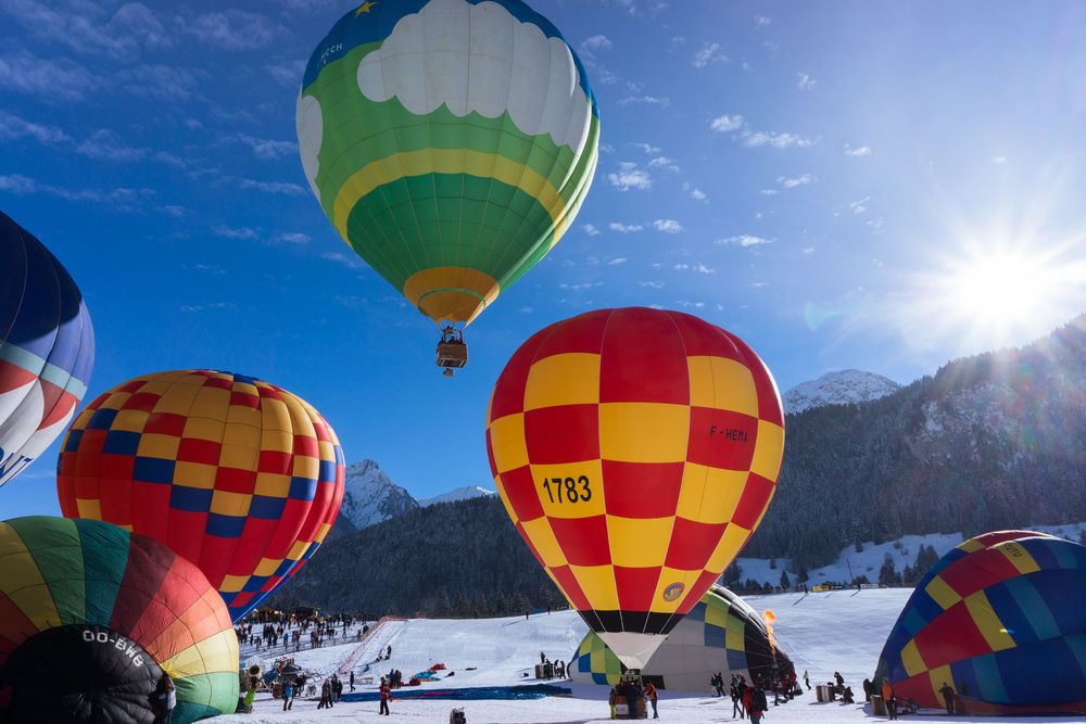 Colorful hot air balloons float above snowy mountains against a bright blue sky during a winter festival.