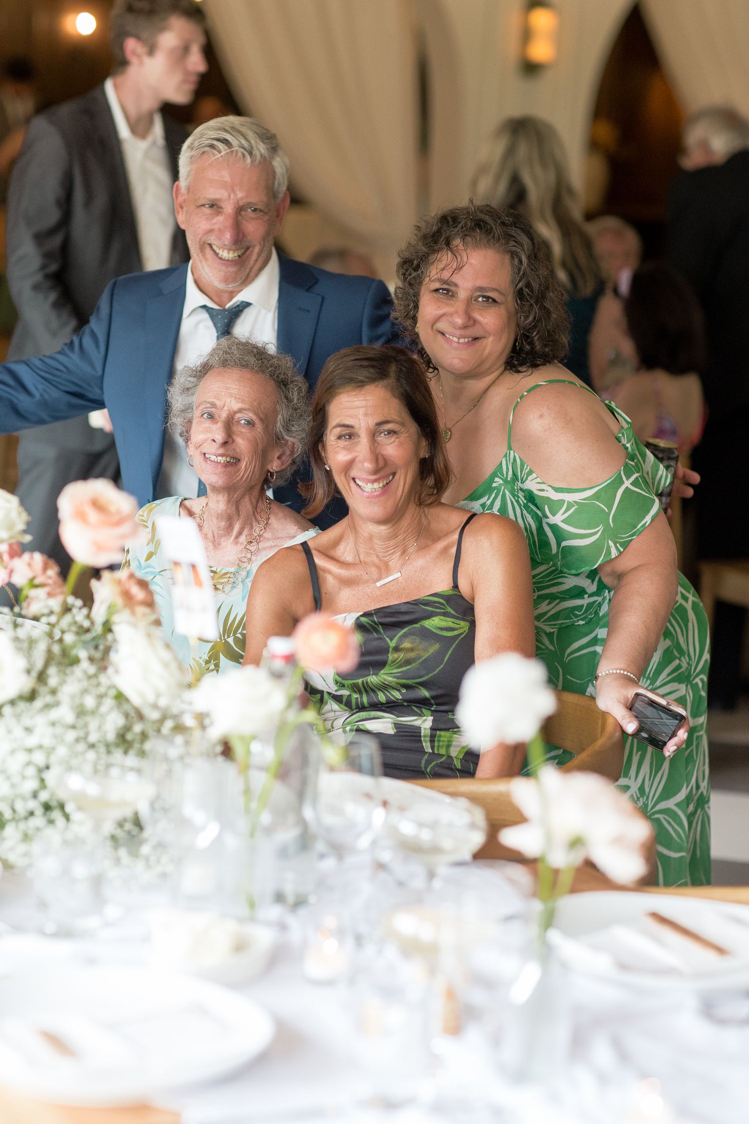 Wedding guests seated at a table decorated with white floral centerpieces share warm smiles during a reception dinner.