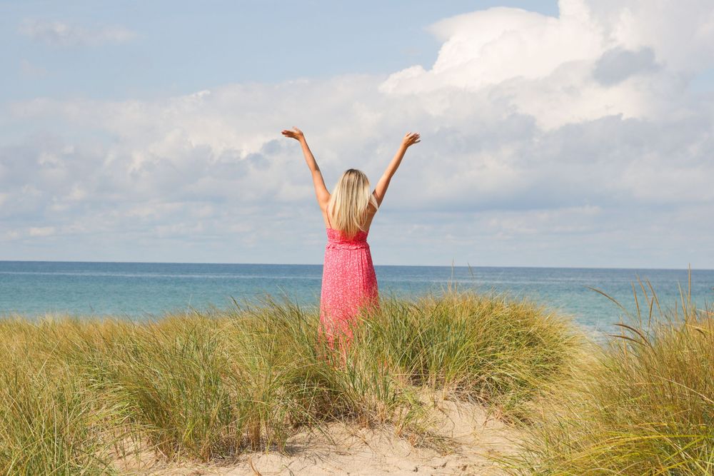 Person in coral dress celebrating on coastal dune with ocean backdrop.