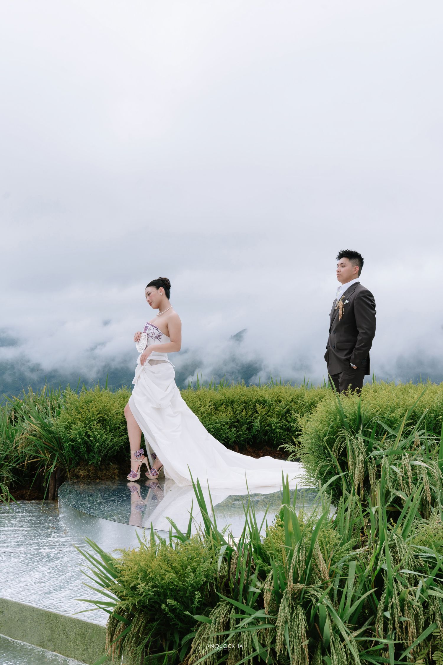 Bride in white gown with train and groom in dark suit posing by water feature in rice fields.
