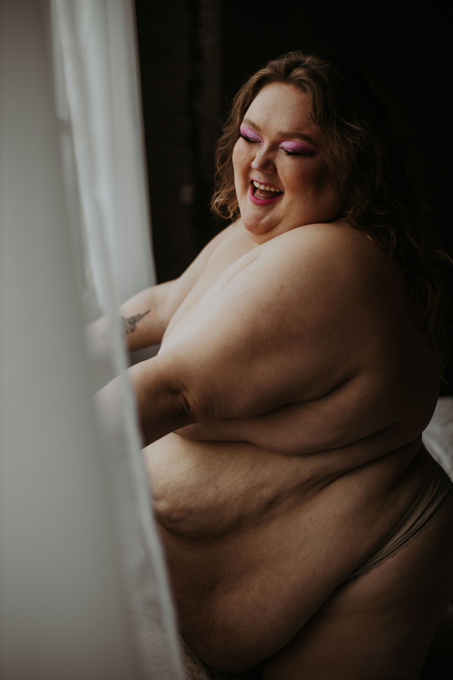 Plus size woman laughing joyfully near a window, wearing minimal clothing, with bold purple eye makeup.
