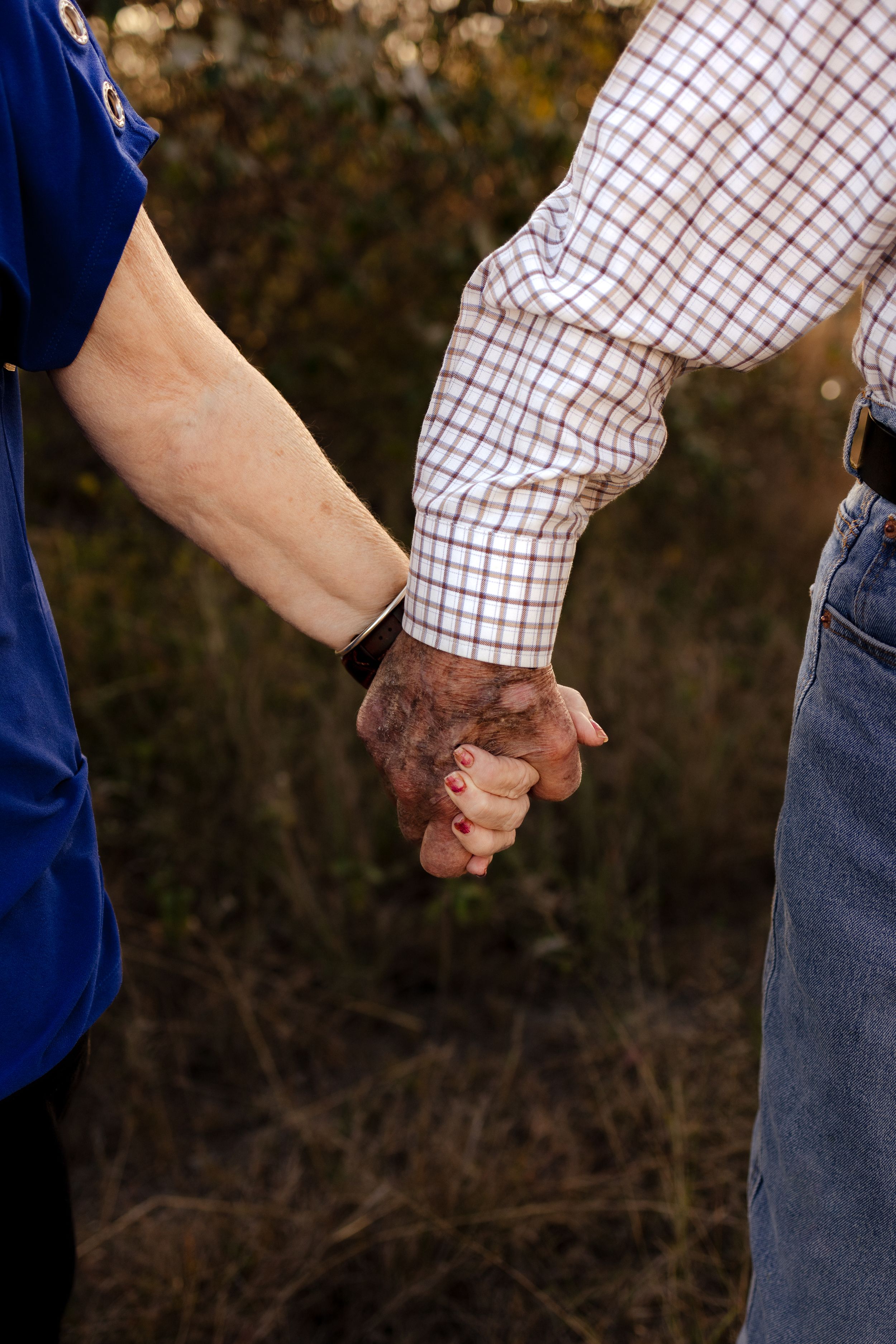 Close-up of elderly couple's hands clasped together showing wedding rings against natural outdoor background.