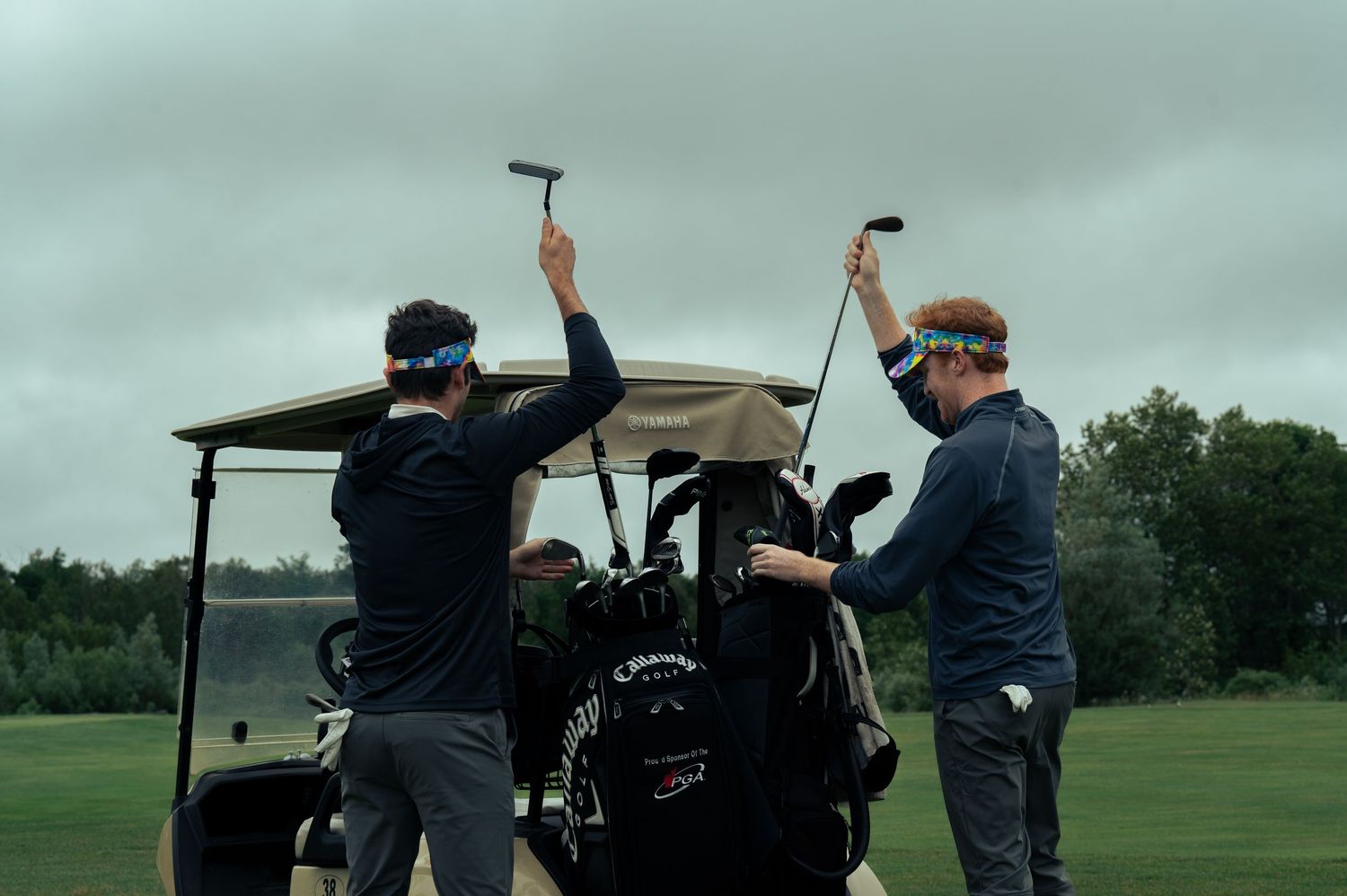 Two golfers raising putters near a Callaway golf bag on a cart on a cloudy golf course.