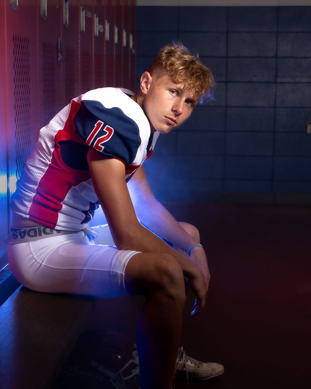 High school football player wearing jersey number 12 sits in locker room with dramatic blue lighting for sports portrait.