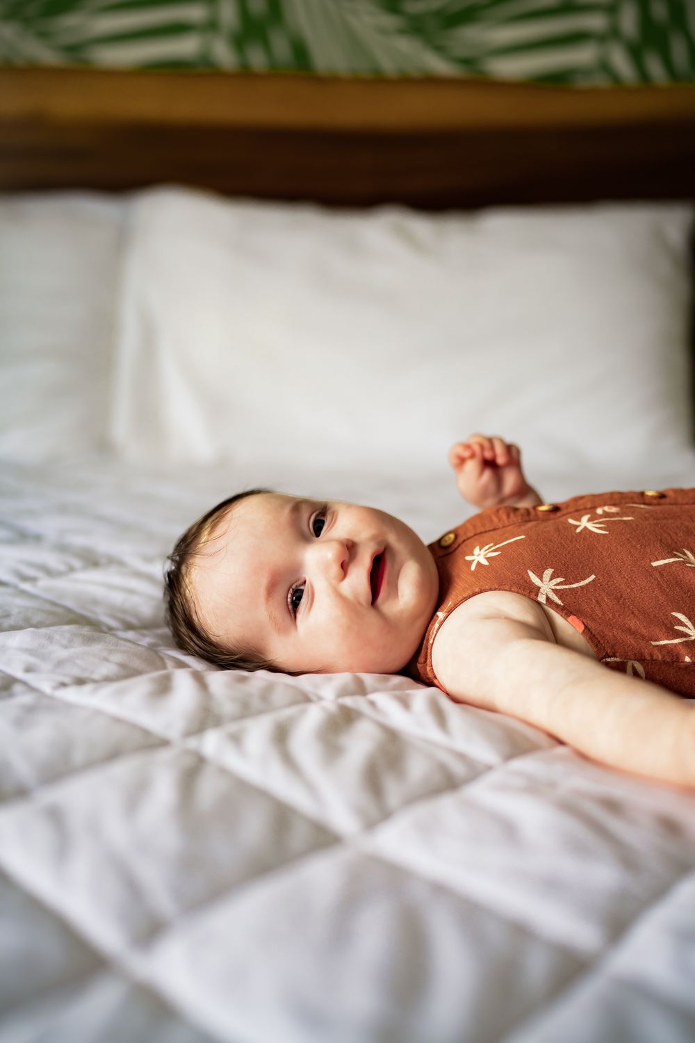 Baby lies on crisp white bedding in a brown onesie looking to the side in a series of connected photos.