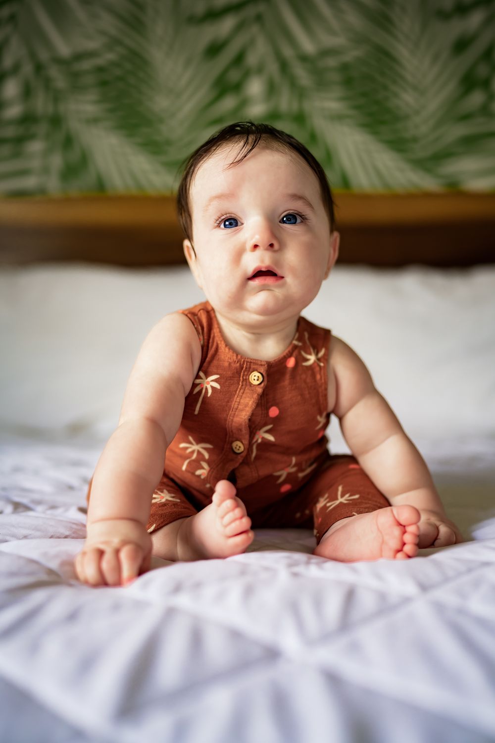 Four sequential photos of a baby in a brown palm tree print outfit lying on white bedding with tropical leaves in background.