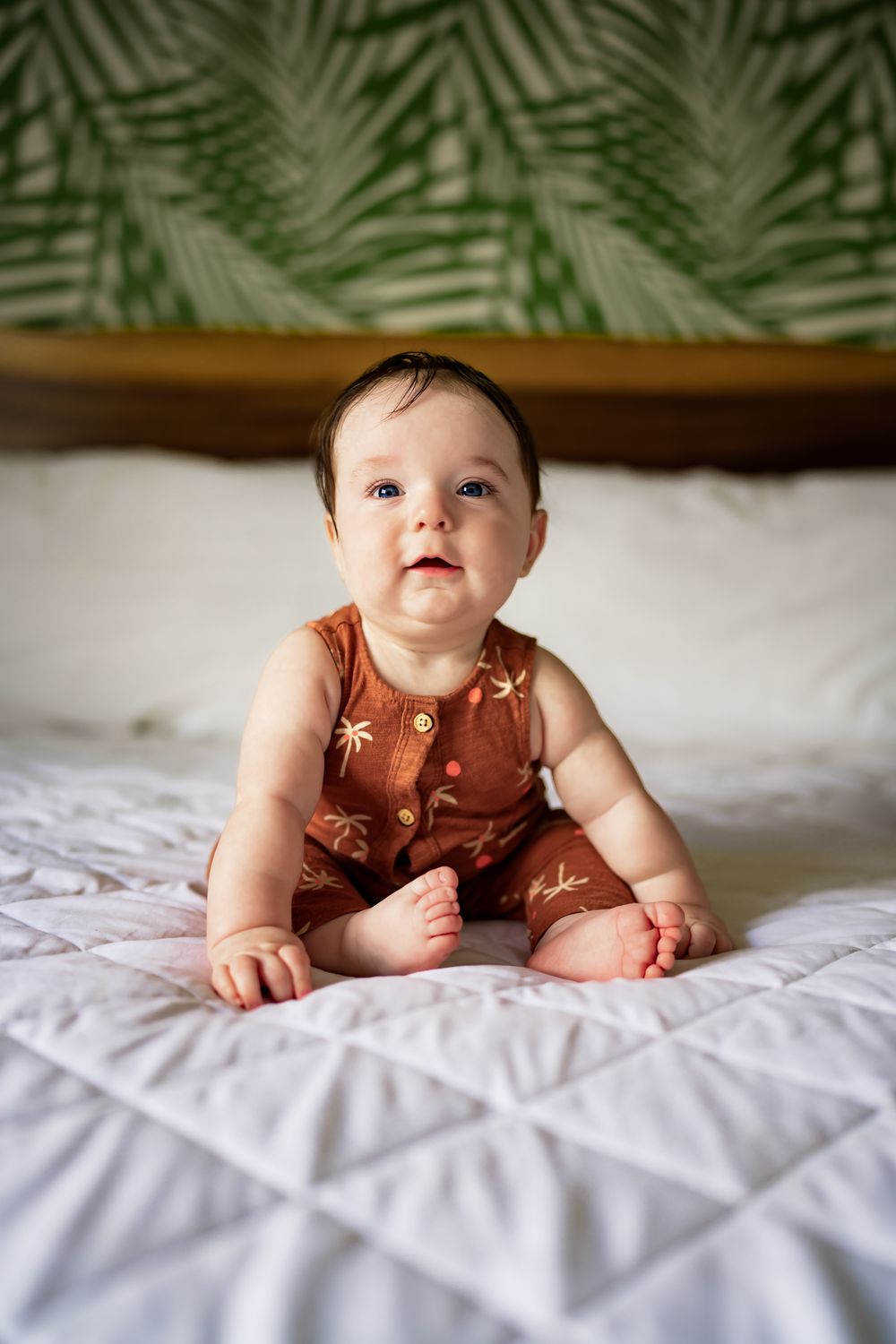Baby wearing brown outfit lies on quilted white bedding with tropical leaves visible in background.