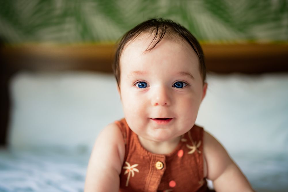 Baby wearing brown outfit lies on white bedding with palm leaf decor in background.