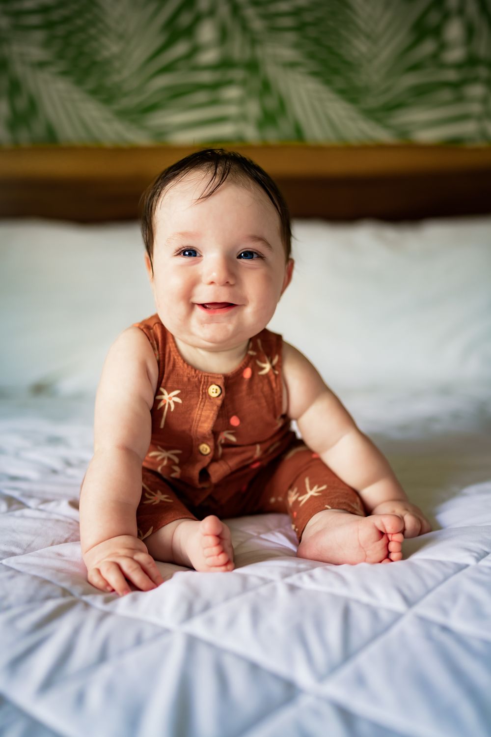 Smiling baby in brown romper lies on white bedding with tropical palm print backdrop.