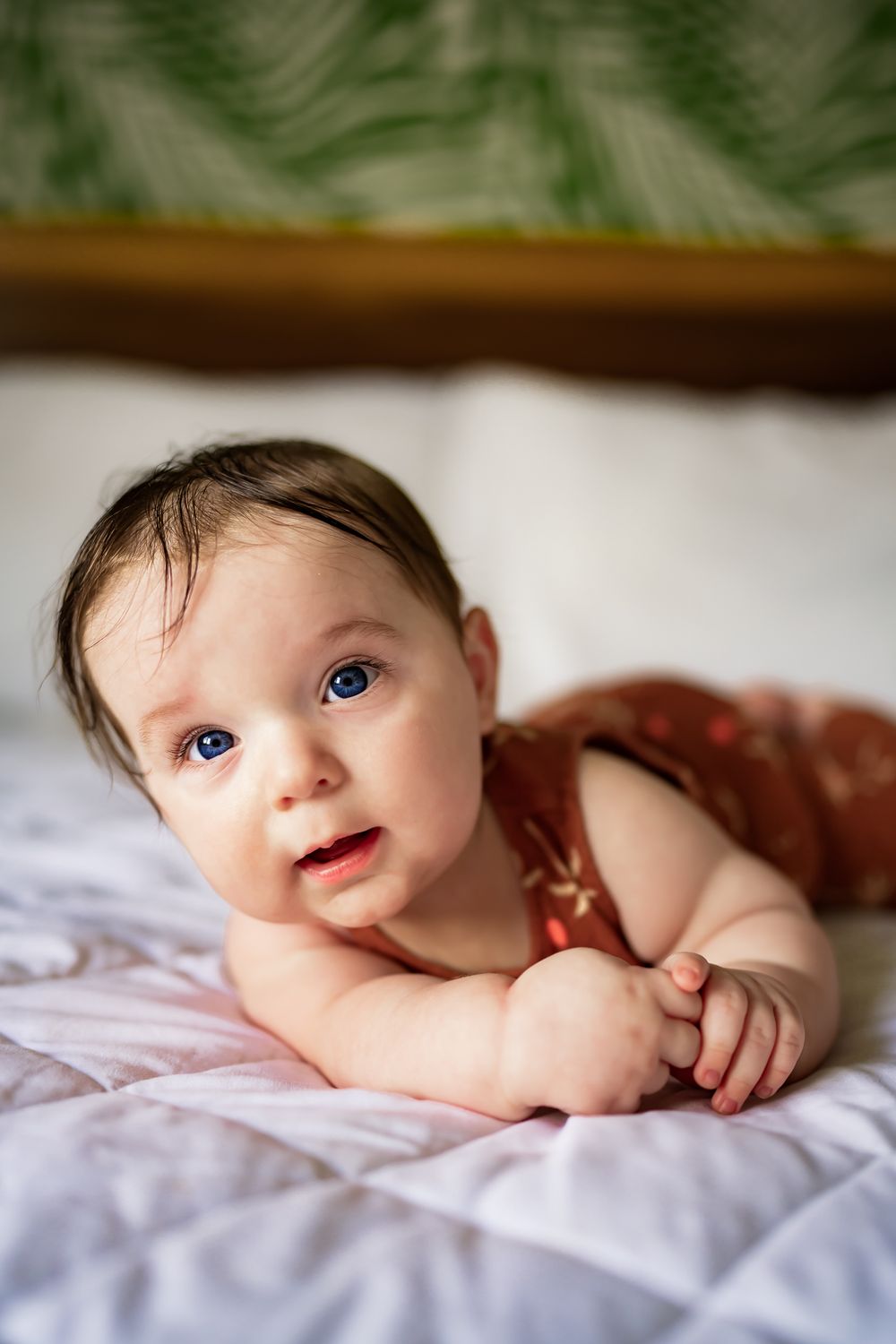 Baby in rust-colored outfit lying on white duvet with green palm fronds decorating the background.