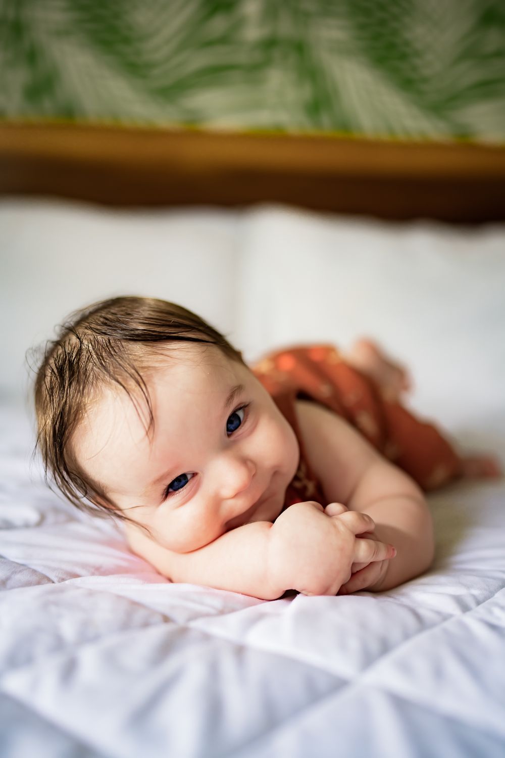 Baby lying on white bedding with blurred green background smiles while resting on tummy.