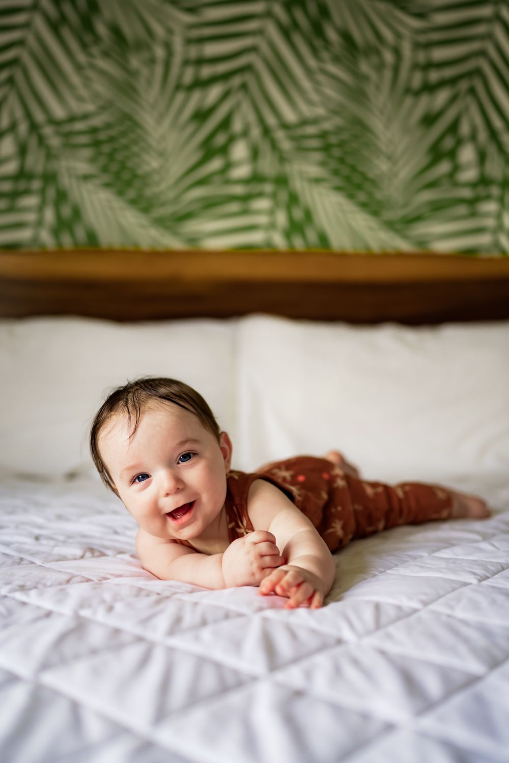 Baby lying on white bedding with whimsical leaf pattern overhead smiles joyfully at camera.