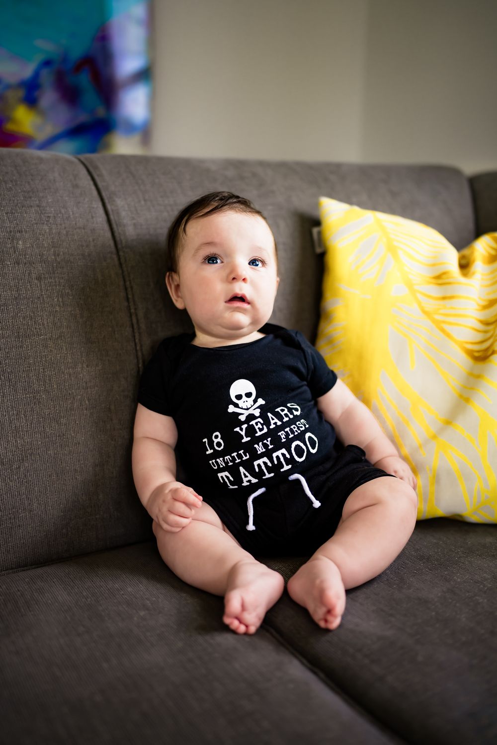 Baby wearing black onesie with skull design sits on grey couch with yellow pillow in background.