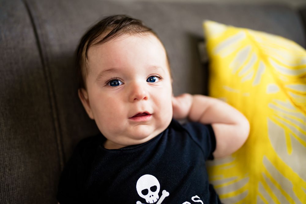 Baby in black skull onesie smiles while sitting on dark grey couch with yellow accent pillow.