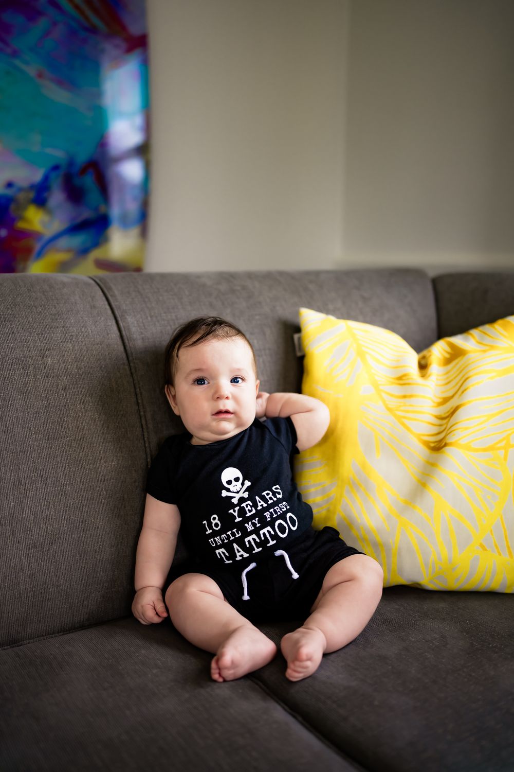 A series of photos showing a baby in a black onesie with text and bone design lounging on a gray couch with yellow pillows.