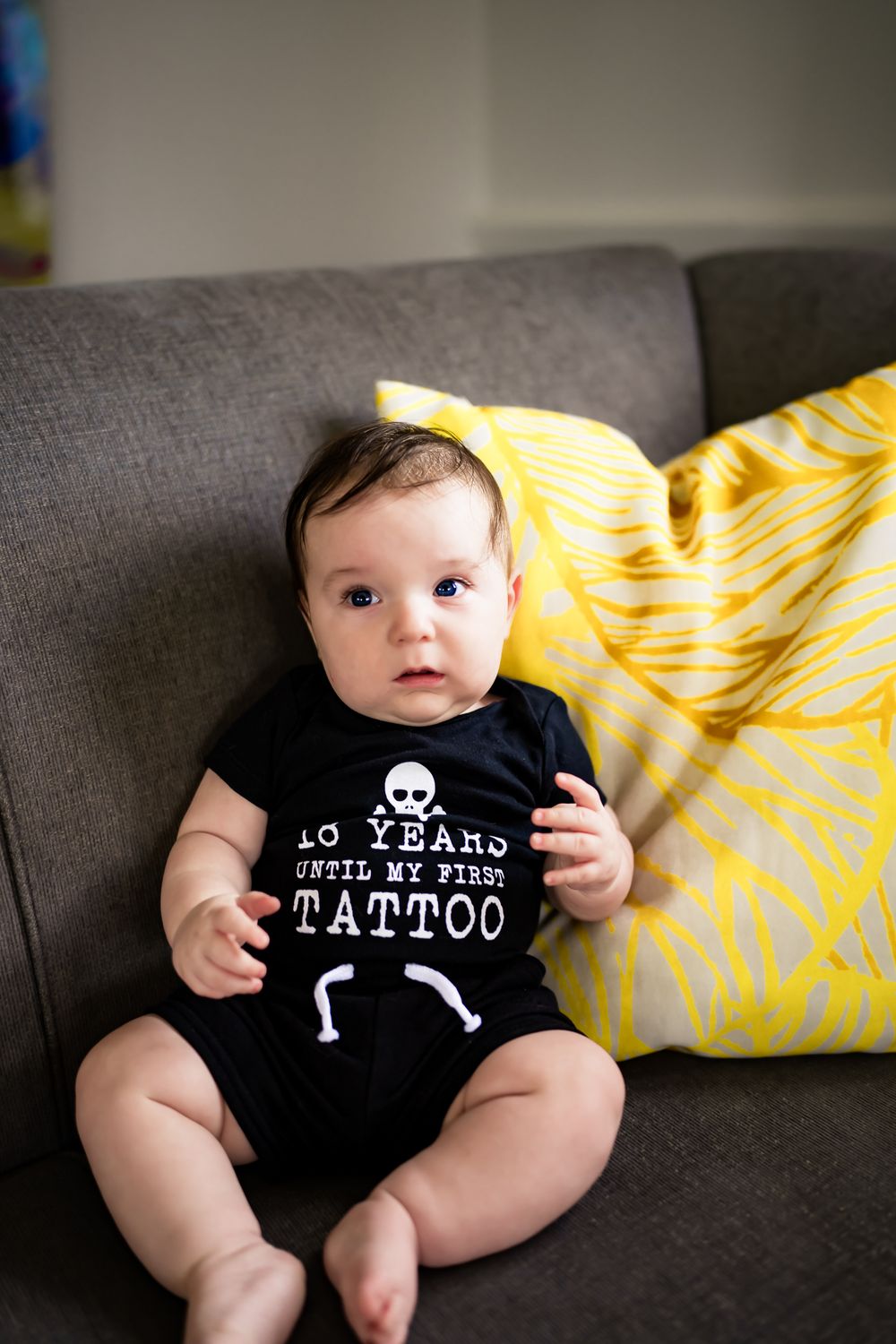Baby wearing black onesie with skeleton design lounges on gray couch with yellow pillow.