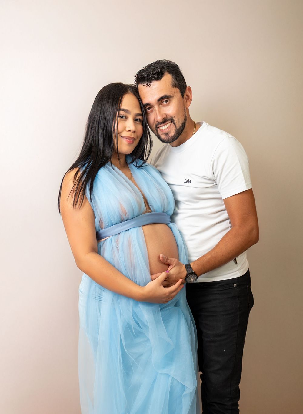 A couple posing together for a maternity photo, with the woman in a flowing blue dress cradling her bump.