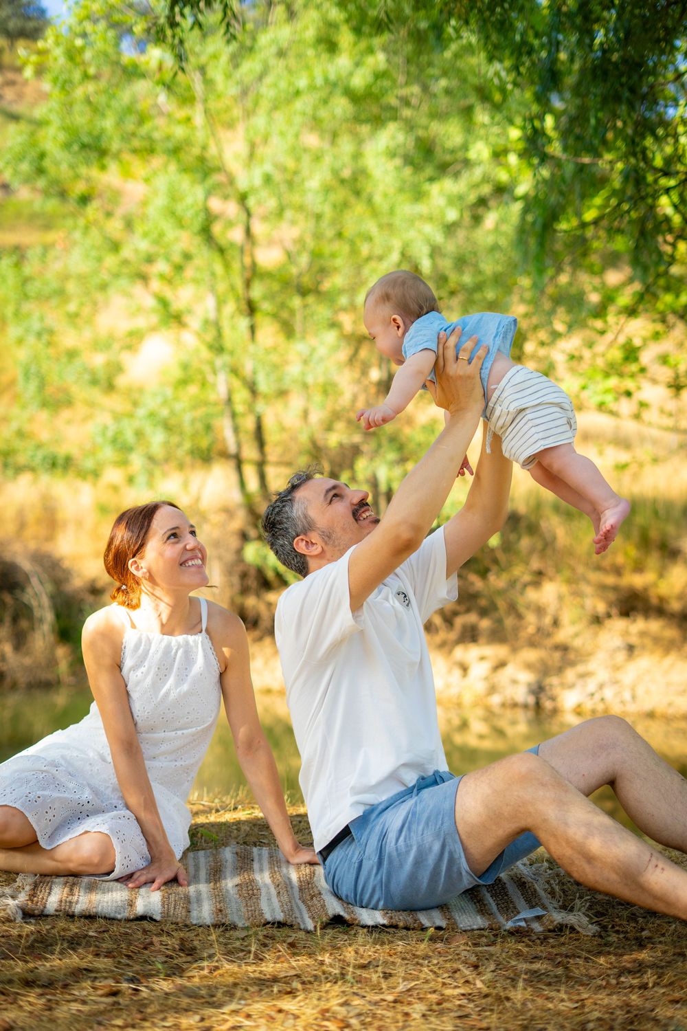 A happy family sits on a blanket outdoors as the father lifts a smiling baby into the air.