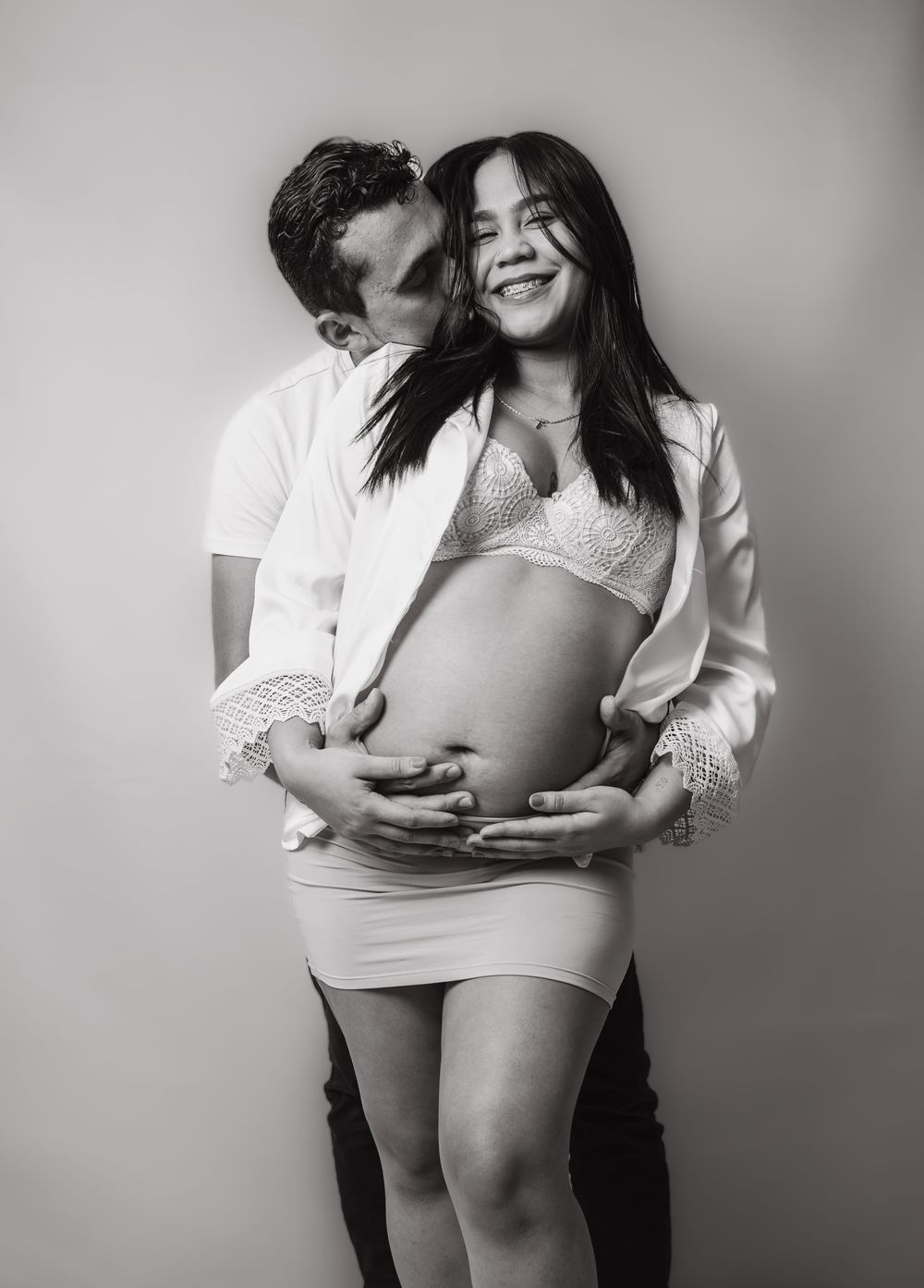 A black and white maternity photo of a smiling pregnant woman being embraced by her partner.