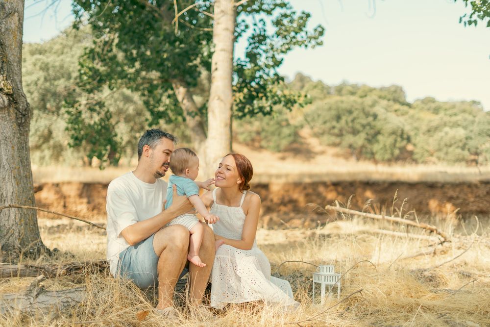A happy family with a baby sits under a tree in a golden field during a warm sunny day.
