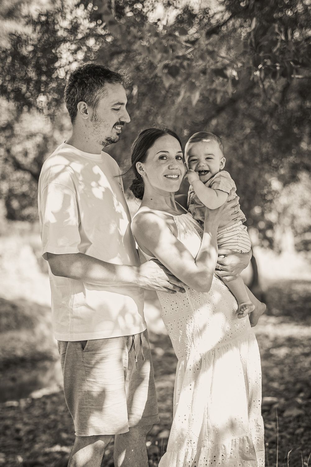A joyful family portrait in sepia tone shows parents holding their laughing baby in a sunlit forest.