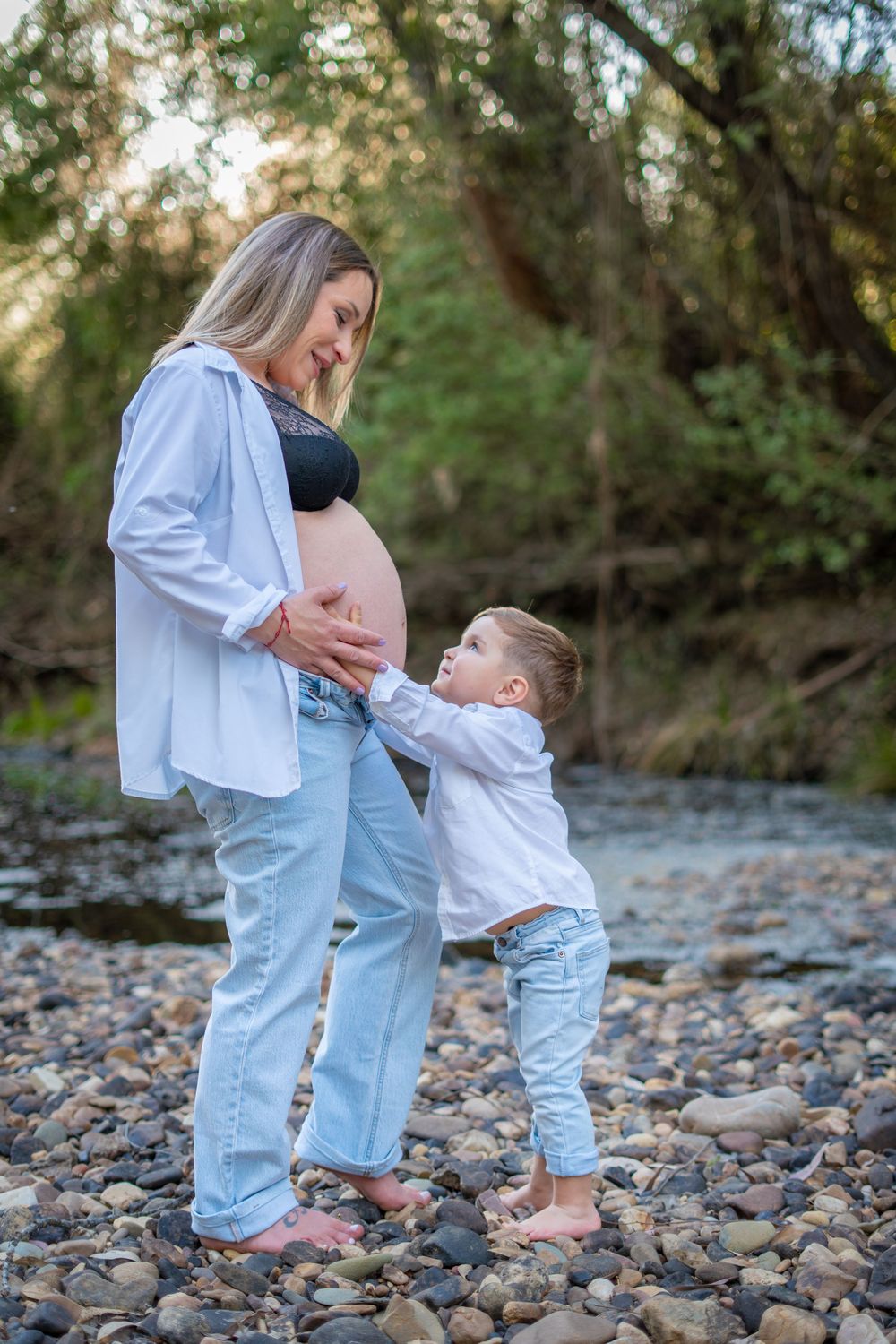 Pregnant woman in jeans and white shirt stands with young child touching her baby bump on rocky riverbank.