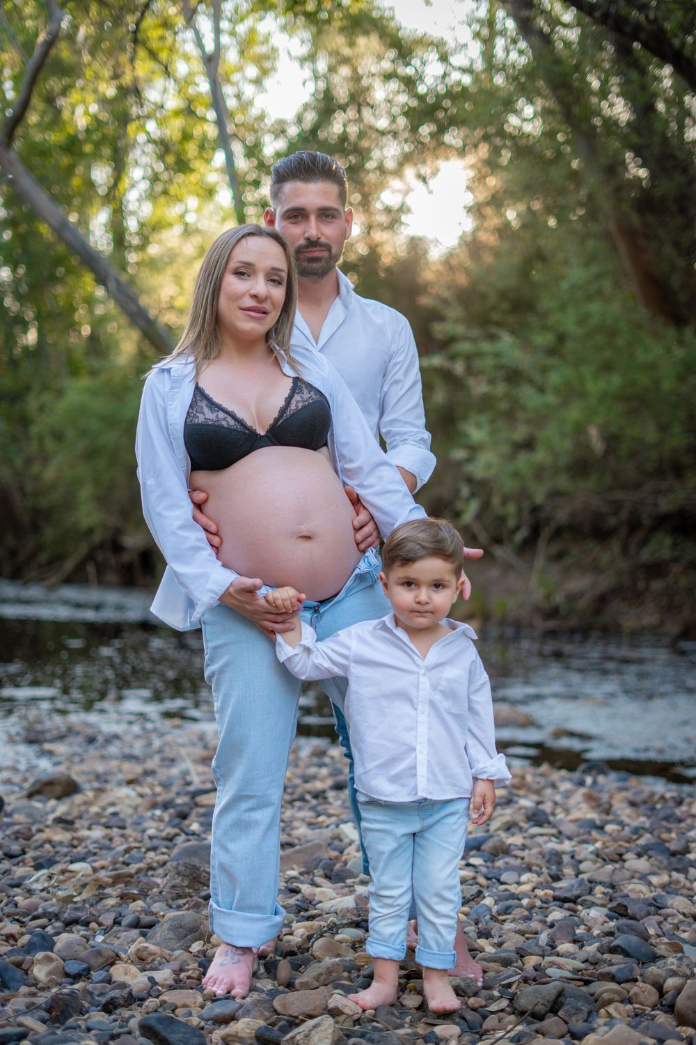 Pregnant woman with partner and child posing outdoors by a rocky creek surrounded by lush green trees at sunset.