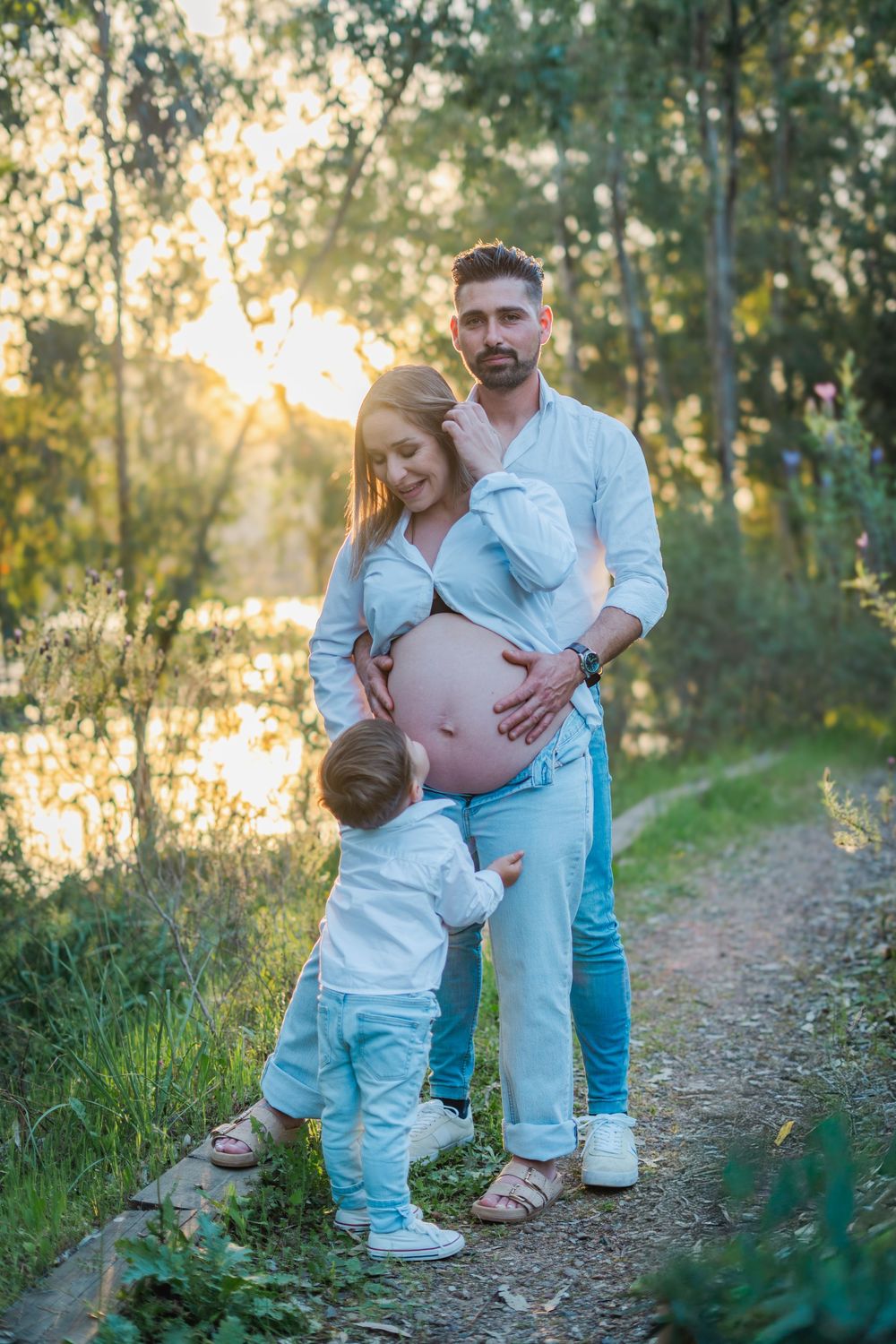 Pregnant woman and man with toddler touching belly on sunlit nature path during golden hour maternity photo session.