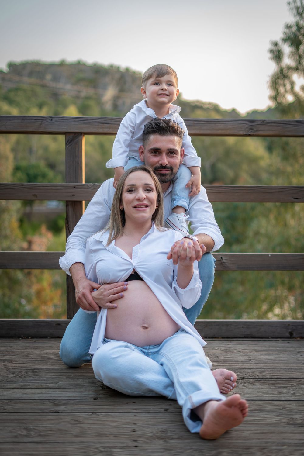 Pregnant woman sits on wooden deck with husband kneeling behind her and young son perched on his shoulders outdoors.