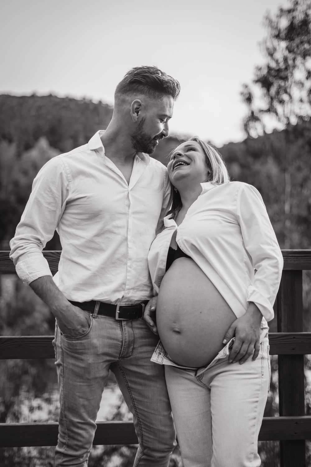 Black and white photo of happy pregnant couple embracing on bridge, woman cradling baby bump.