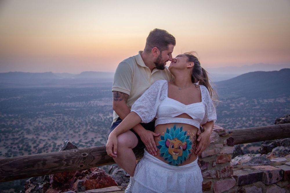 A couple shares a tender kiss at sunset on a hilltop, showcasing a colorful pregnancy belly painting.