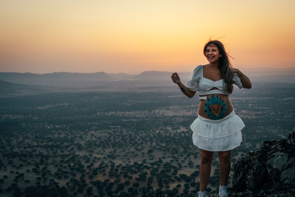 A pregnant woman with belly art poses on a hilltop at sunset with a vast landscape behind her.
