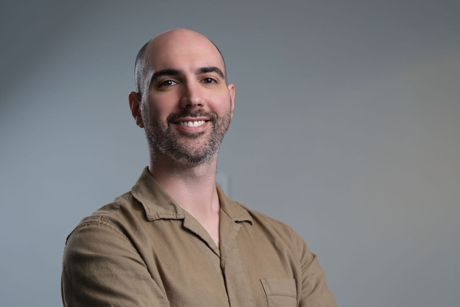 Smiling bald man with beard poses with crossed arms in casual tan shirt for professional headshot. Iron Galazy Studios.