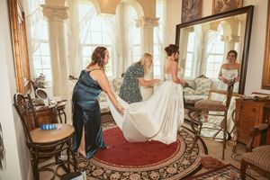 Wedding party helping bride with dress preparations in an elegant Victorian-style room with ornate furniture and windows.