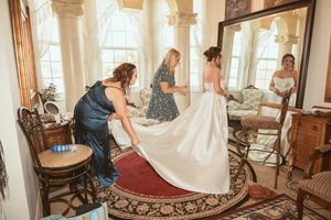 Wedding party assisting bride with dress and preparations in a sunlit Victorian room with vintage furnishings.