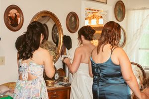 Women in formal dresses arrange their hair while looking into vintage oval mirrors mounted on a wall.