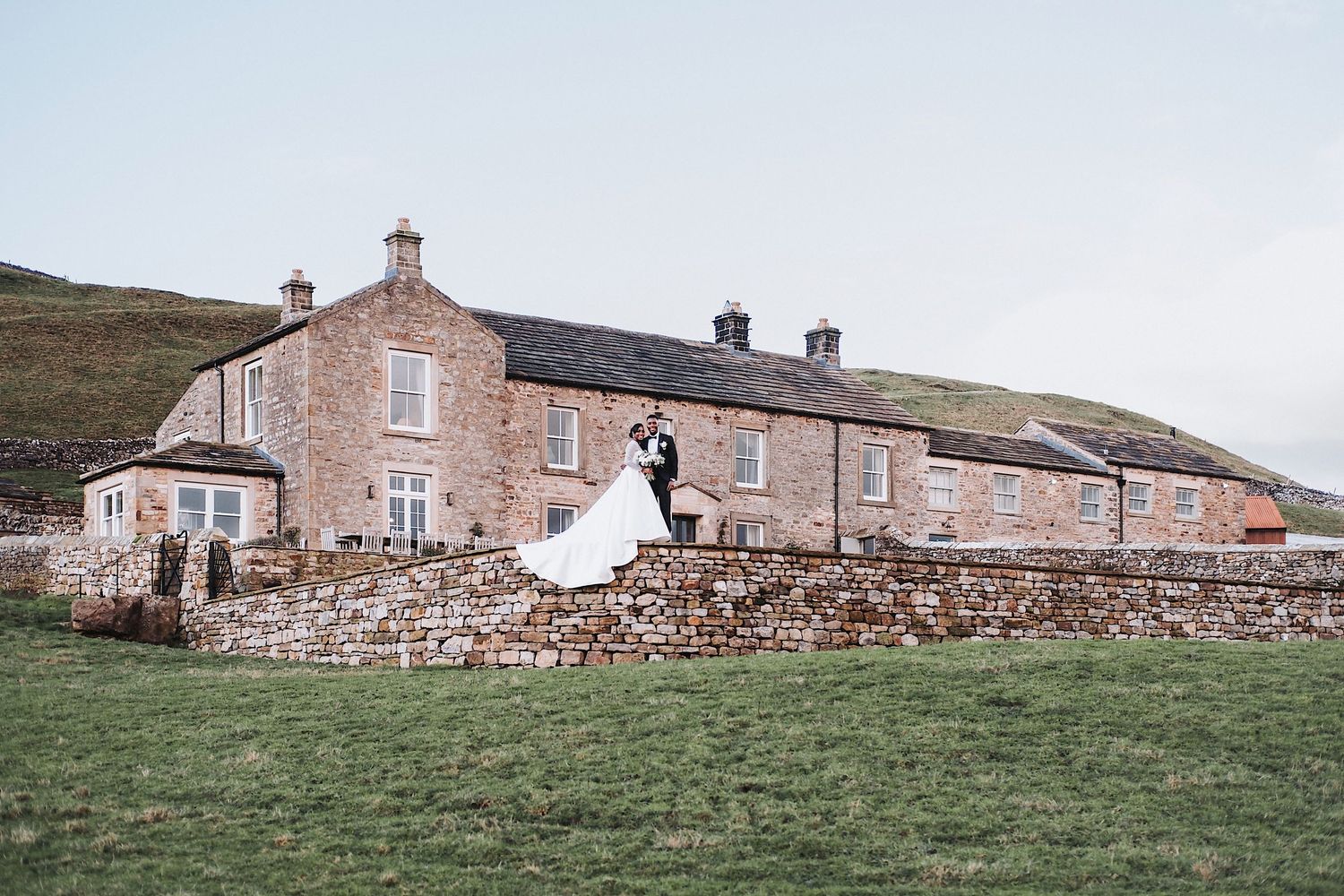 Rustic stone cottage buildings stretch along green countryside hill with church steeple visible.