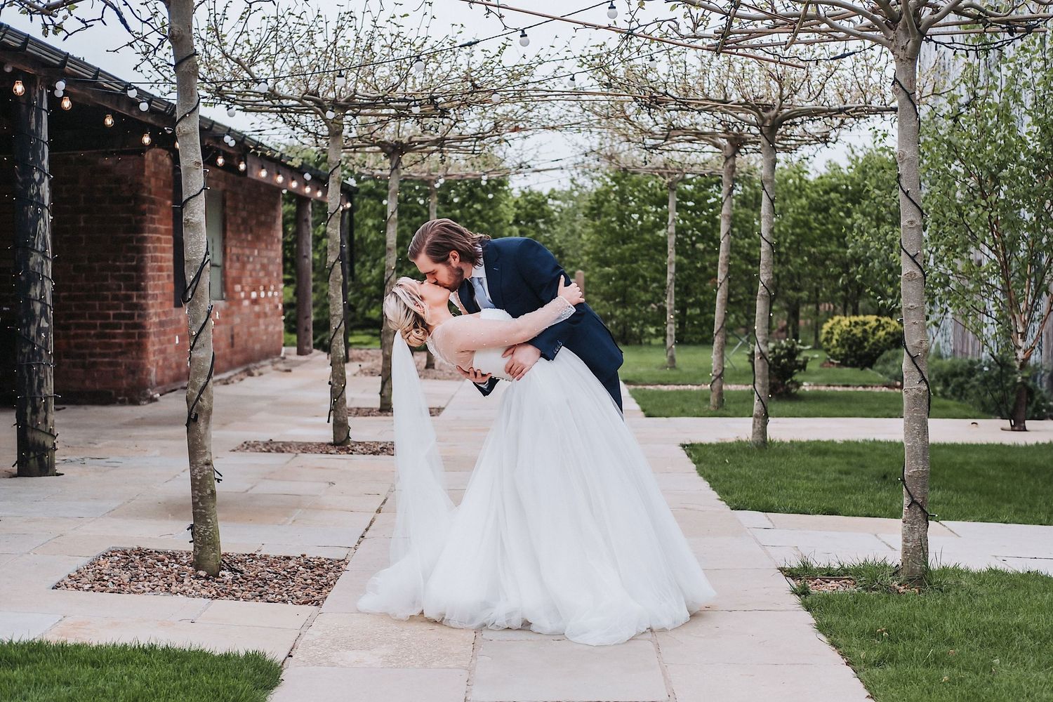 Elegant outdoor wedding scene with bridesmaids in lavender dresses and a bride in white at a venue with brick walls and trees.