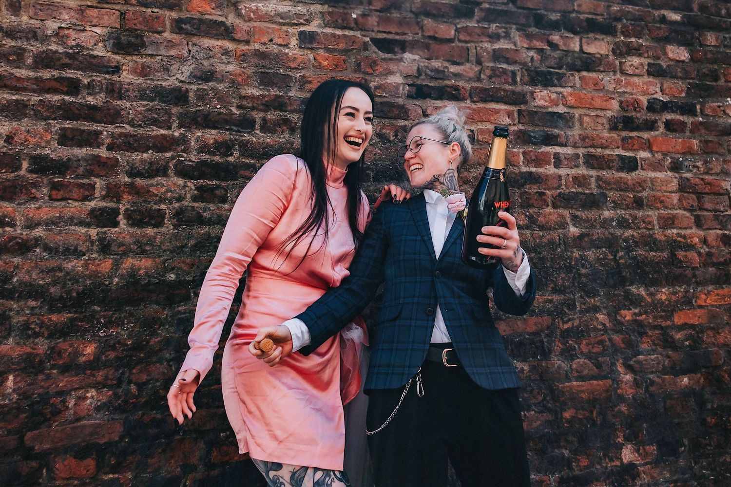 Two friends in pink dress and blue blazer laugh together while drinking champagne against brick wall.